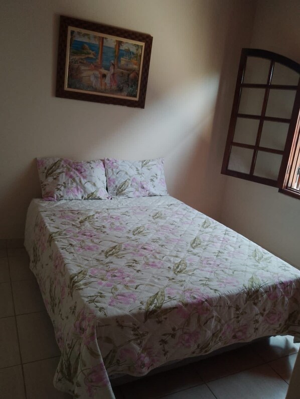 A bedroom with a bed, floral bedspread, a painting on the wall, and a window with a wooden frame.