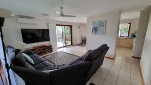 A living room with a black sofa, a flat-screen TV, a ceiling fan, and a dining area with a table and chairs. - Welcome to "Seascapes" (Gympie)
