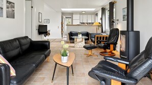 Ein modernes Wohnzimmer mit einem schwarzen Ledersofa, einem Holztisch und einem schwarzen Ledersessel. In der Ecke ist ein schwarzer Kaminofen zu sehen.