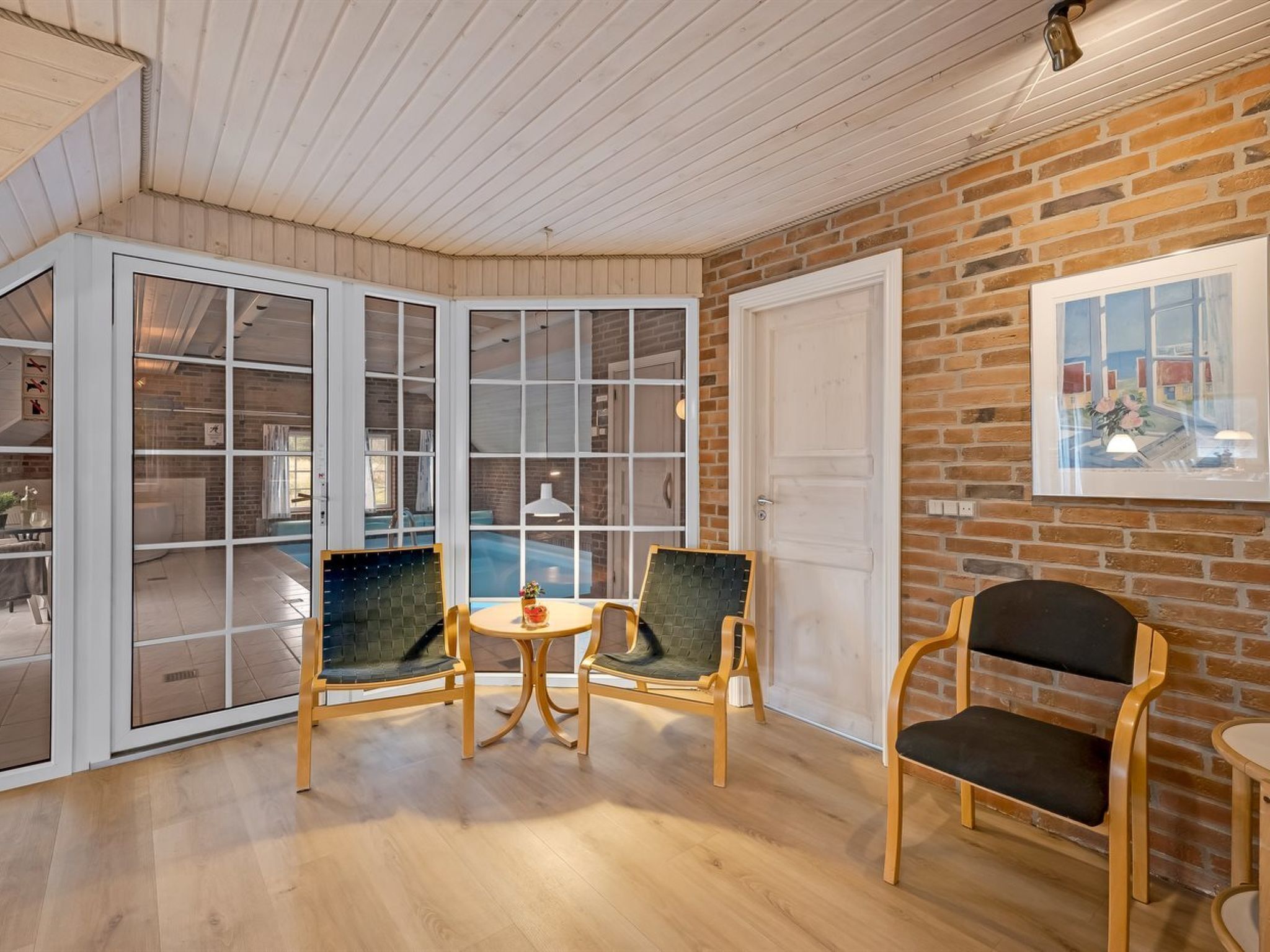 A room with a wooden floor, two green upholstered chairs around a small round table, a white door, and a brick wall with a framed picture.