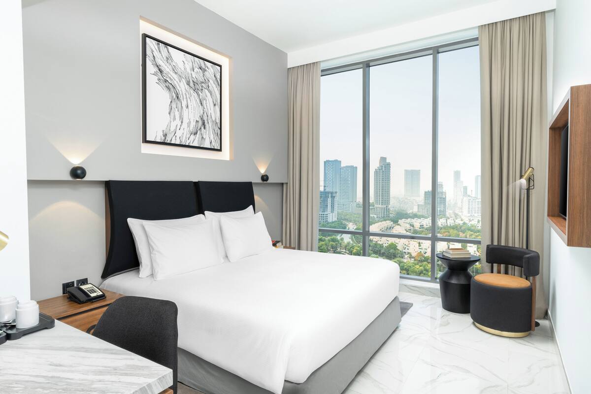 A modern hotel room with a large bed, a city view, a desk with a phone, and a small table with books.