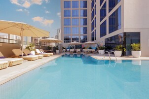 A rooftop pool with lounge chairs and umbrellas, surrounded by a modern building with large windows.