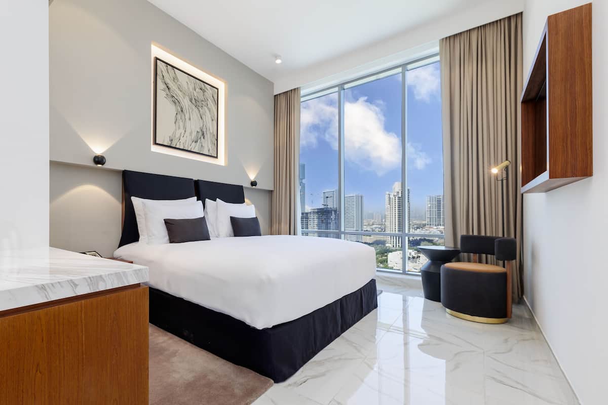 A modern hotel room with a large bed, a desk, a chair, and a view of the city skyline.