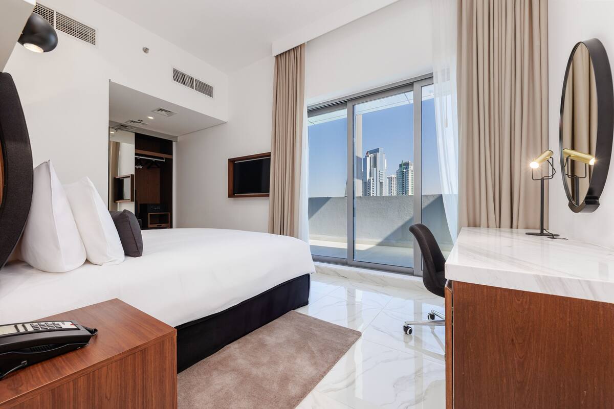 A modern hotel room with a large bed, a desk, a chair, a TV, and a view of the city through large windows.