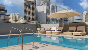 Une piscine sur le toit, dotée de chaises longues et d’un parasol, entourée d’immeubles de grande hauteur.