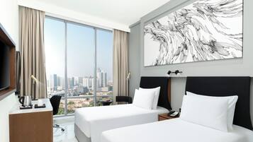 Quarto de hotel com duas camas, uma escrivaninha, uma cadeira, uma janela grande com vista para a cidade e um quadro na parede.