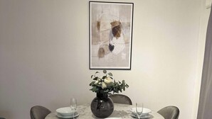 Een eetgedeelte met een ronde tafel, vier stoelen, een vaas met bloemen en een lijst met een abstract schilderij aan de muur.