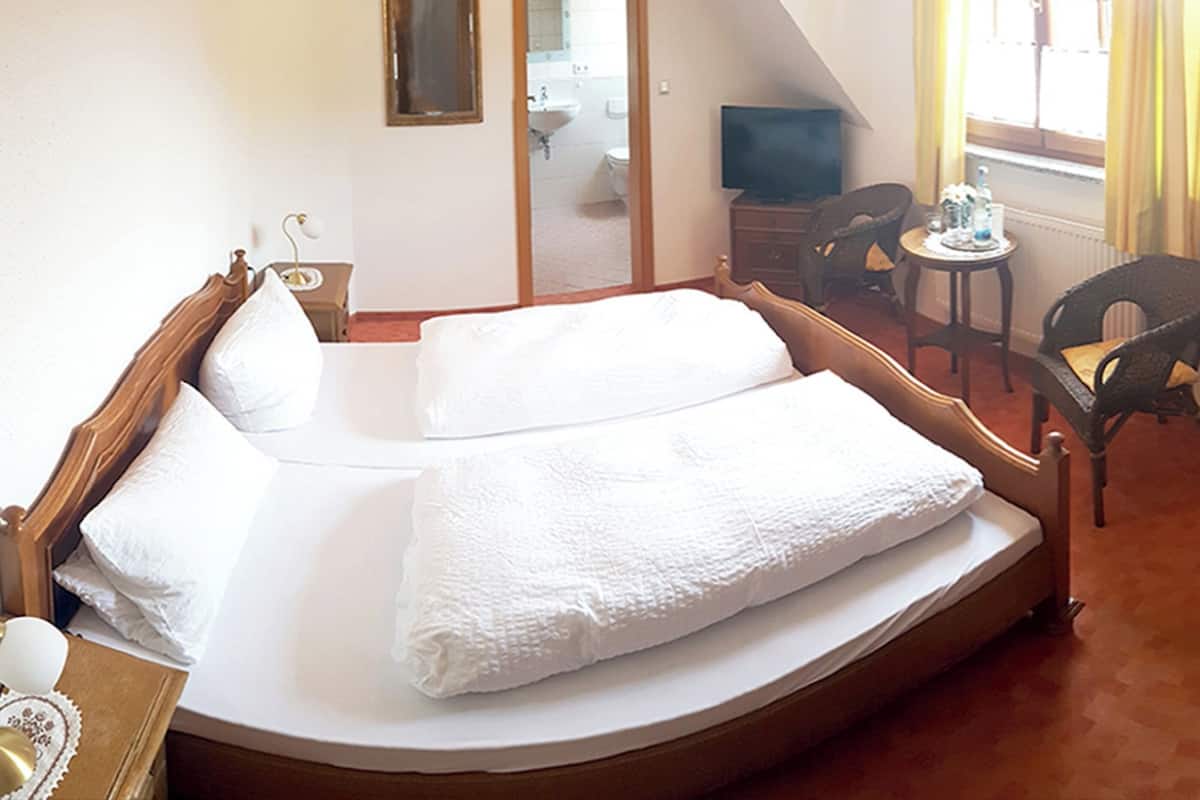 A bedroom with a single bed, a small table, two chairs, a television, and a bathroom visible through an open door.