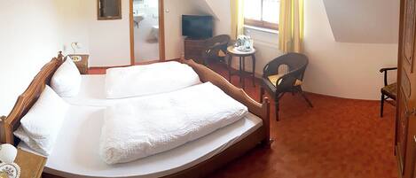 A bedroom with a single bed, a small table, two chairs, a television, and a bathroom visible through an open door.