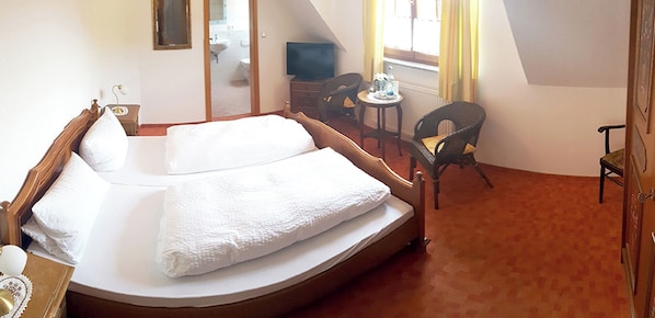 Un dormitorio con una cama individual, una mesita, dos sillas, un televisor y un baño visible a través de una puerta abierta.