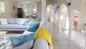 Uma sala de estar moderna com um sofá cinzento, almofadas azuis e amarelas, uma mesa de centro e uma área de jantar ao fundo.