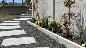 Uma trilha conduz a um edifício com uma parede de concreto cinzento e um jardim com diversas plantas.