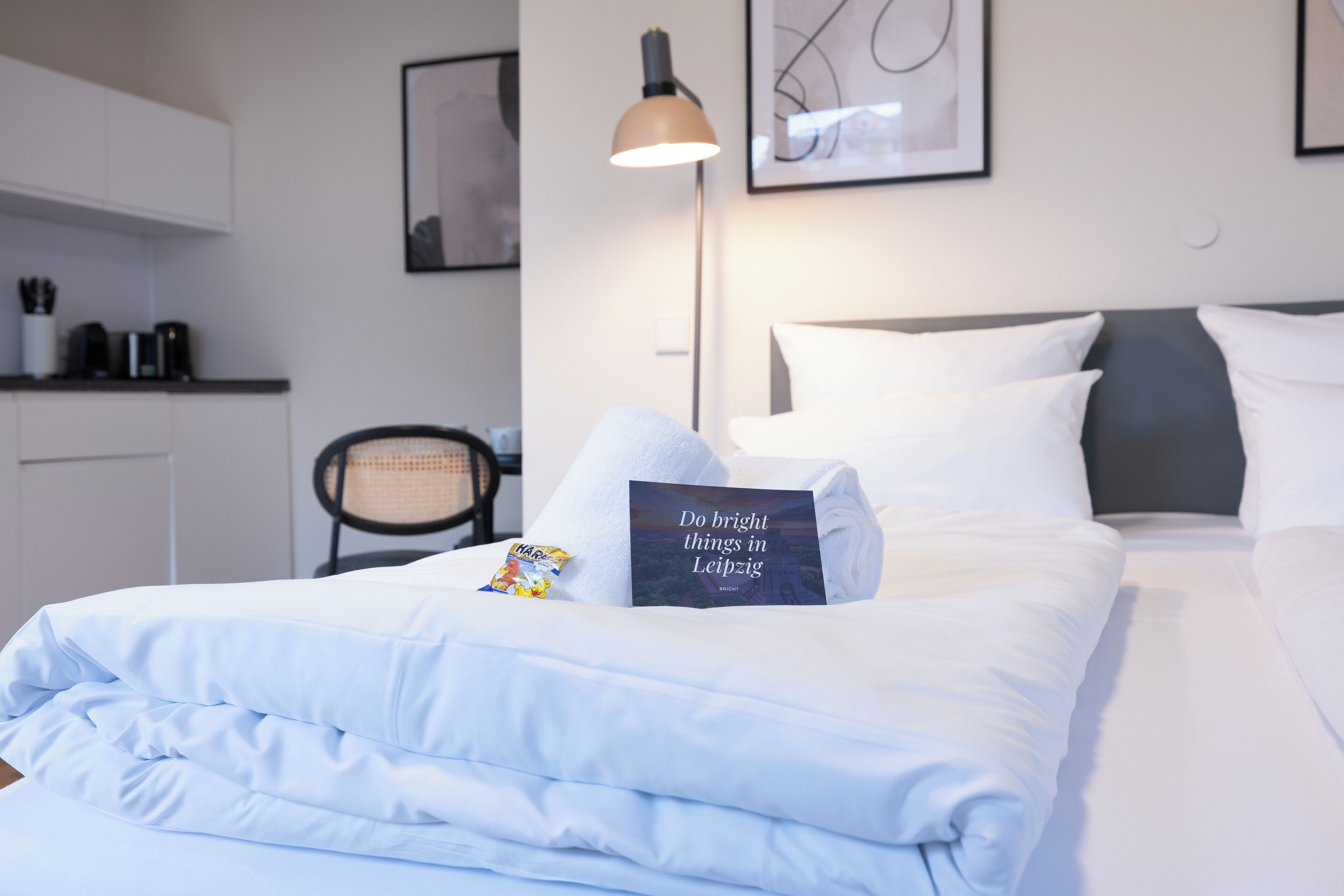 Una cama bien hecha con sábanas blancas, una almohada con texto, una lámpara y un cuadro en la pared.