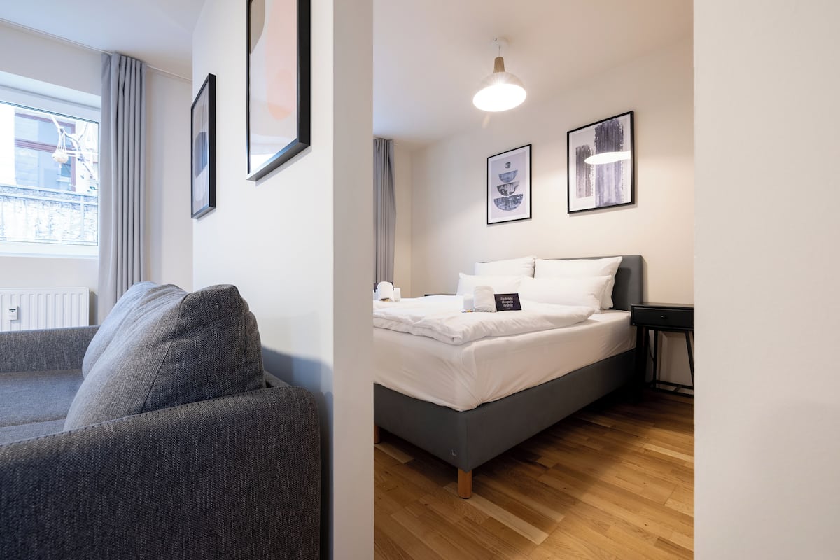 A modern hotel room with a grey sofa, a neatly made bed with white linens, and a wooden floor.