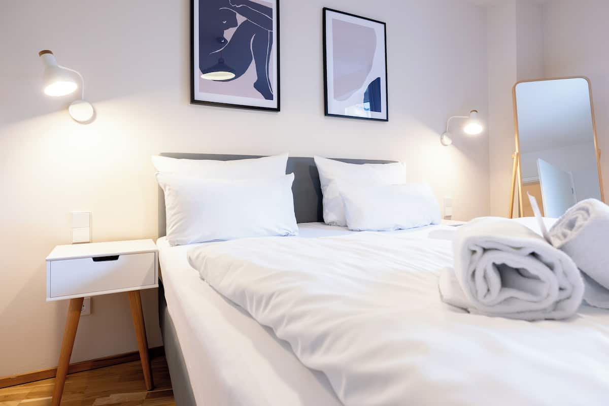 A neatly made bed with white linens, a white bedside table with a drawer, and two wall-mounted light fixtures.