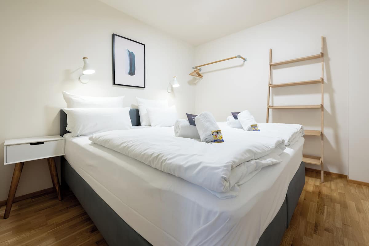 A neatly made bed with white linens, a bedside table with a drawer, a wall-mounted light, a framed picture, and a wooden ladder against the wall.