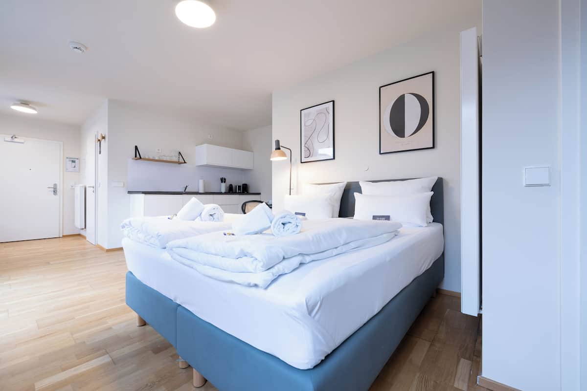 A modern hotel room with a neatly made bed, a kitchenette, and a bathroom visible in the background.
