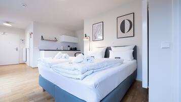 A modern hotel room with a neatly made bed, a kitchenette, and a bathroom visible in the background.