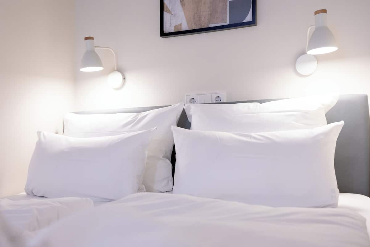 A neatly made bed with white linens and pillows, two wall-mounted reading lamps, and a framed picture above the bed.