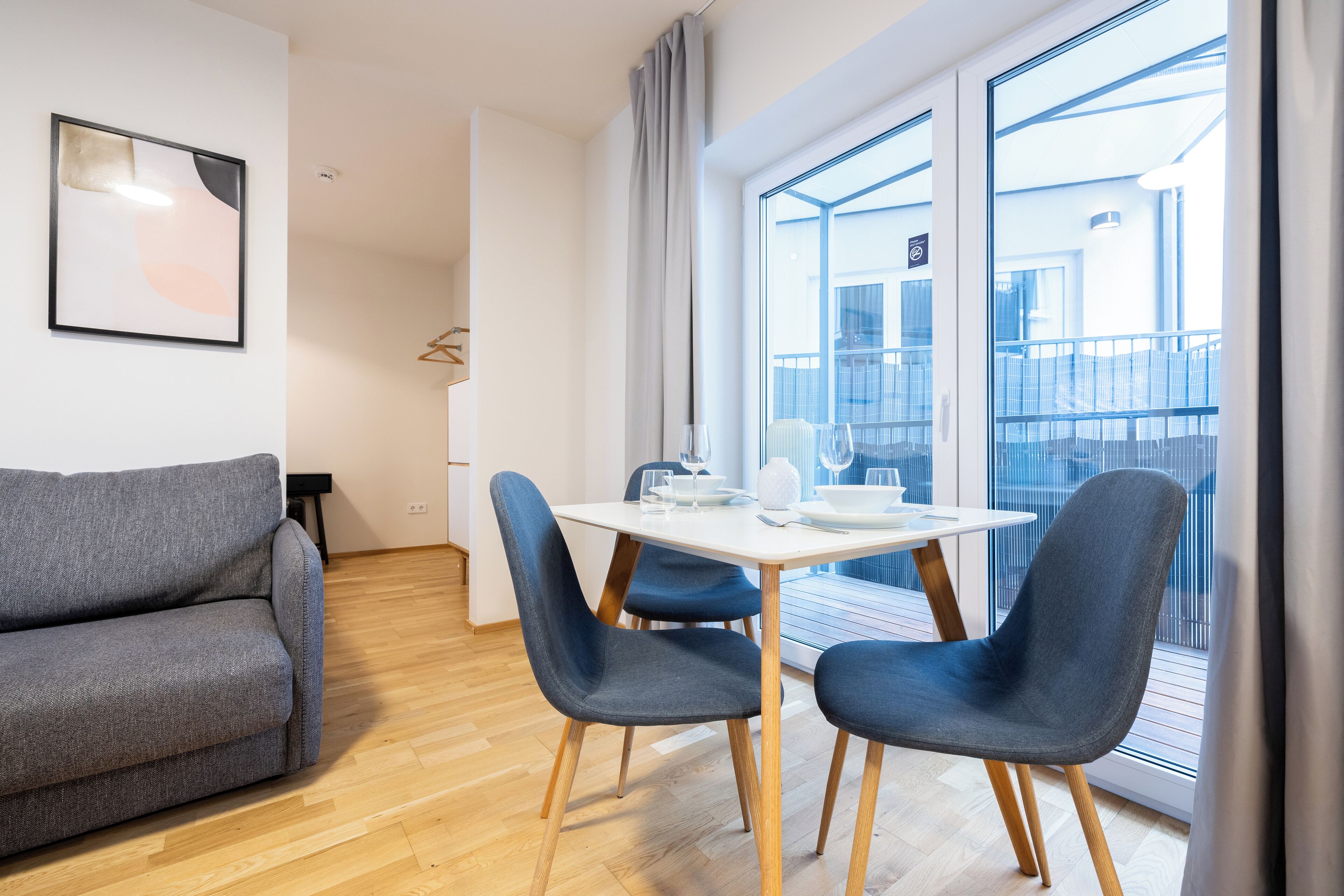A modern living room with a grey sofa, a dining table with chairs, and a large window with a view of the city.