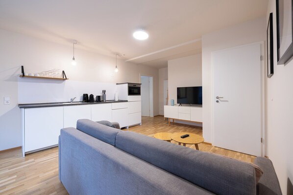 A modern apartment with a grey sofa, a small wooden table, a kitchen with white cabinets, and a TV on a white stand. - BRIGHT Leipzig Zentrum-West Stadium (Leipzig)