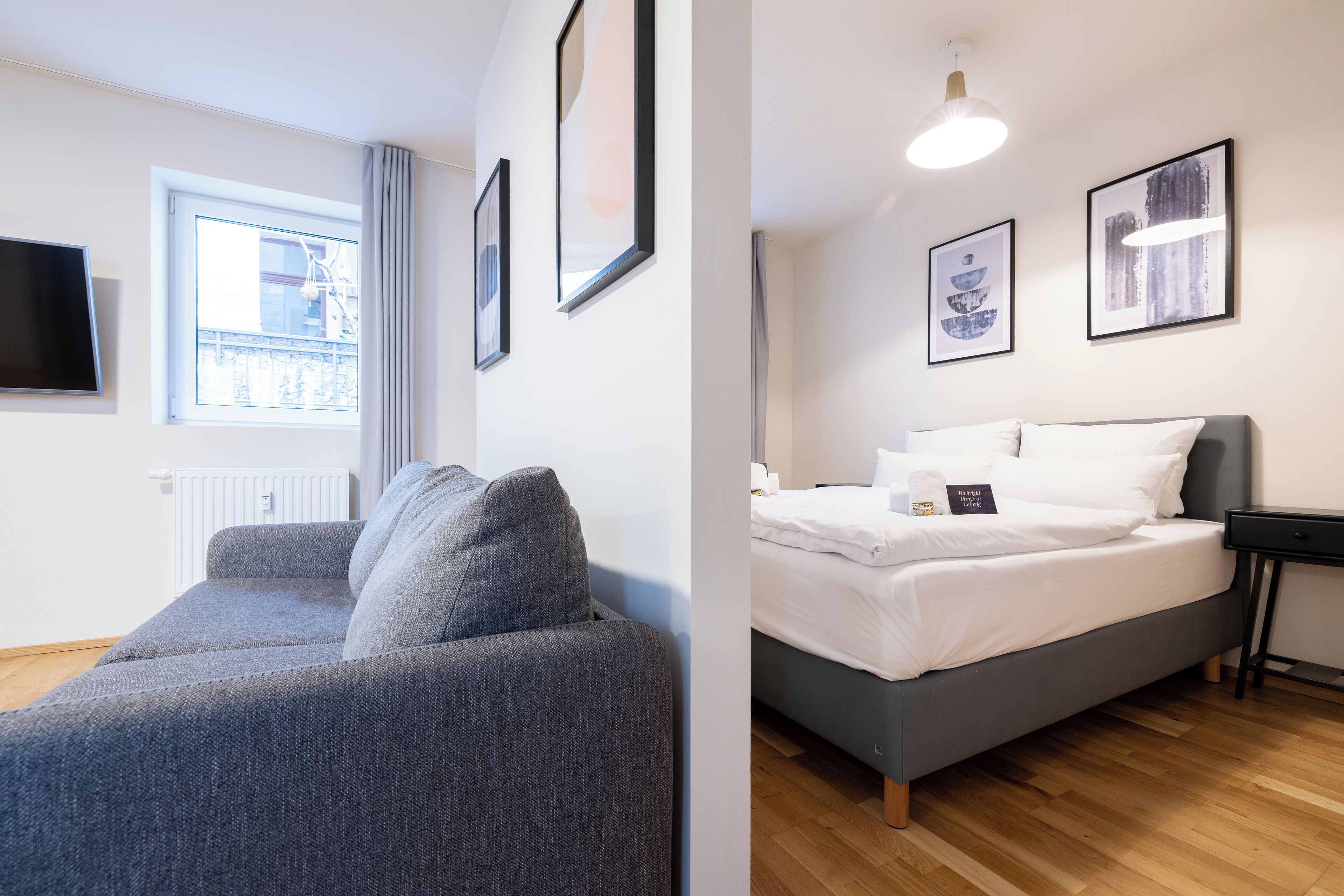 A modern room with a grey sofa, a bed with white bedding, a bedside table, and framed artwork on the wall.