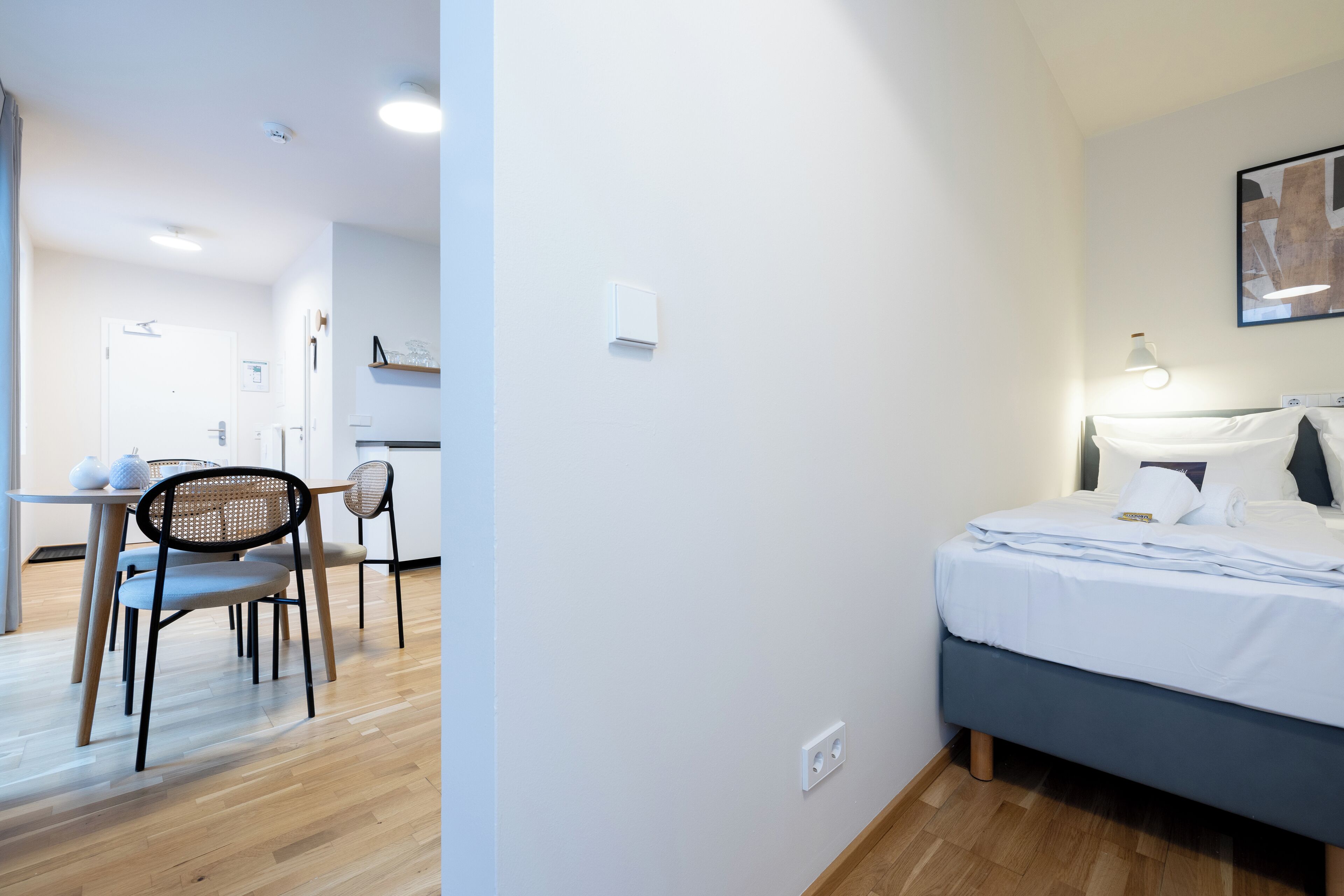A modern hotel room with a bed, bedside table, and a dining area with chairs and a table.