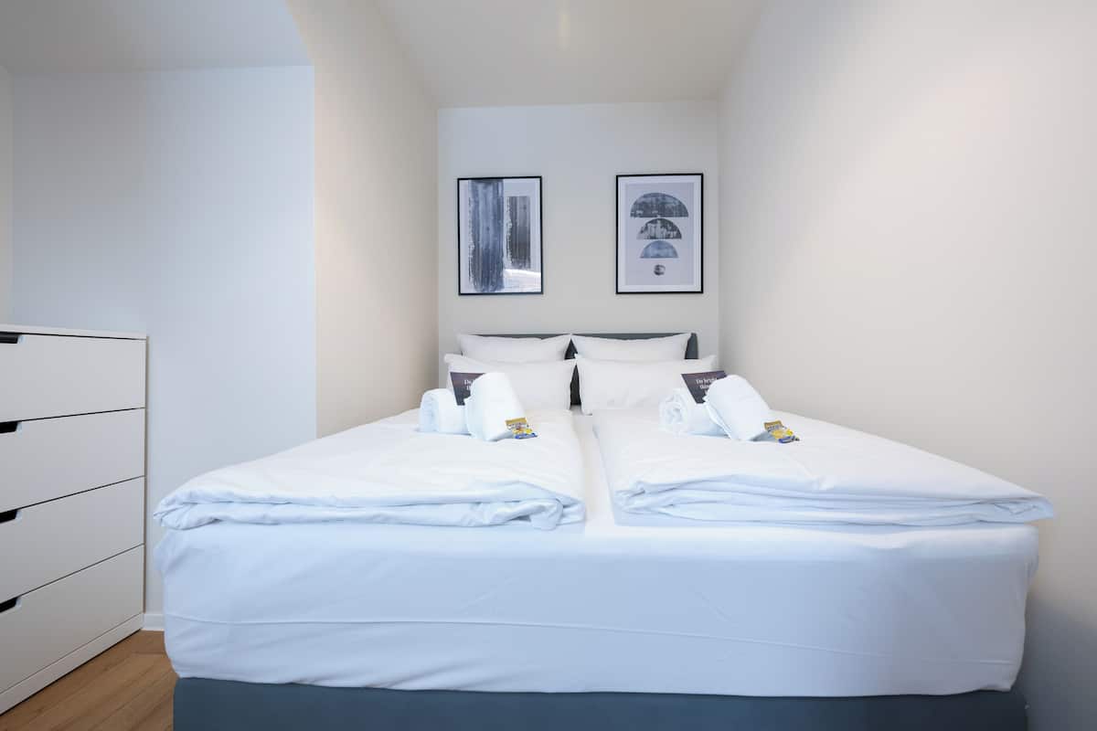 A neatly made twin bed with white linens, a bedside table with a lamp, and two framed artworks on the wall.