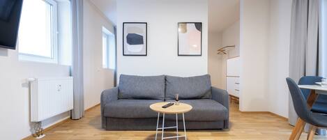A modern living room with a grey sofa, a round wooden coffee table, and a flat-screen TV mounted on the wall.