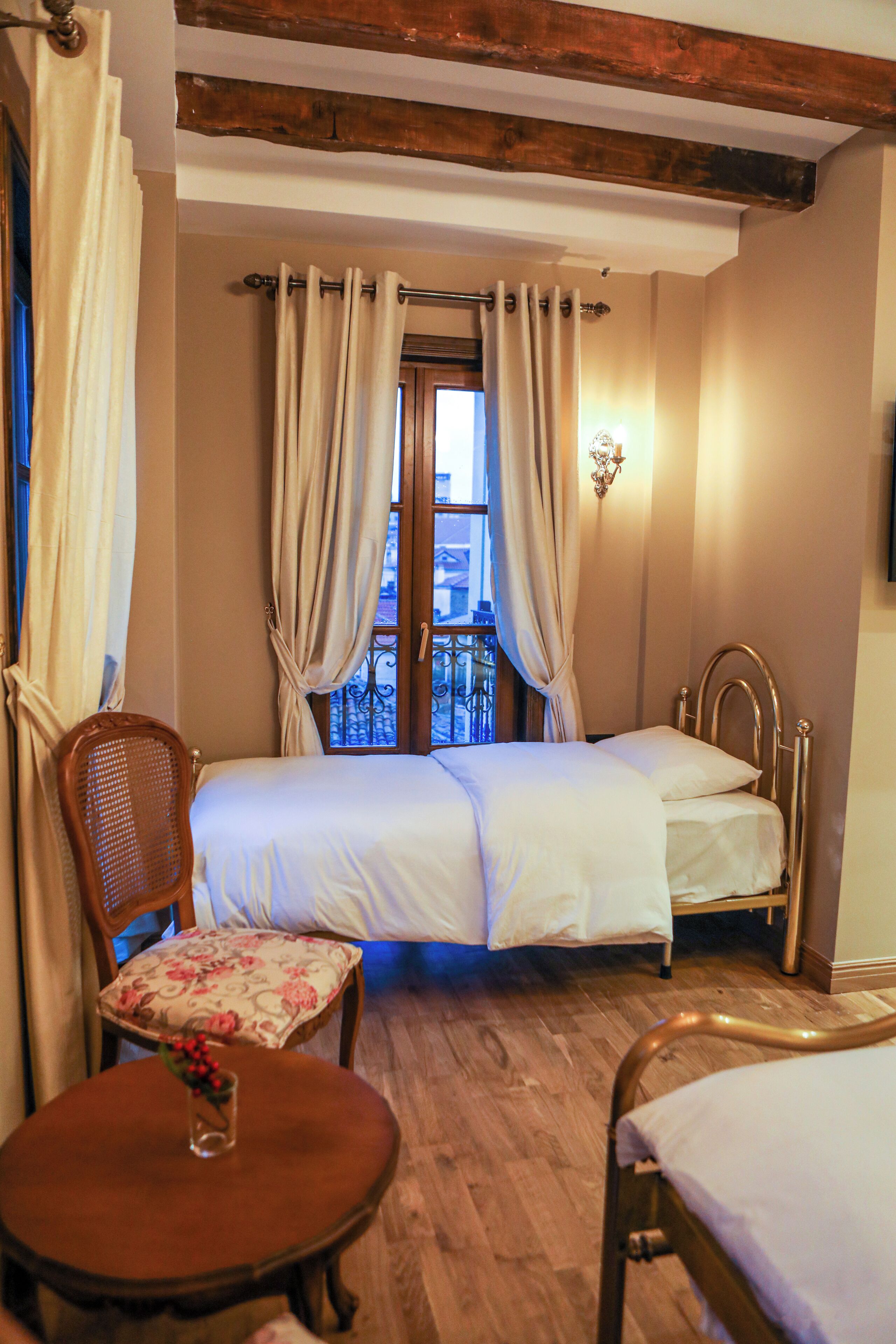 Une chambre à coucher avec un lit double, une chaise en bois, une petite table avec un vase et une fenêtre avec des rideaux.