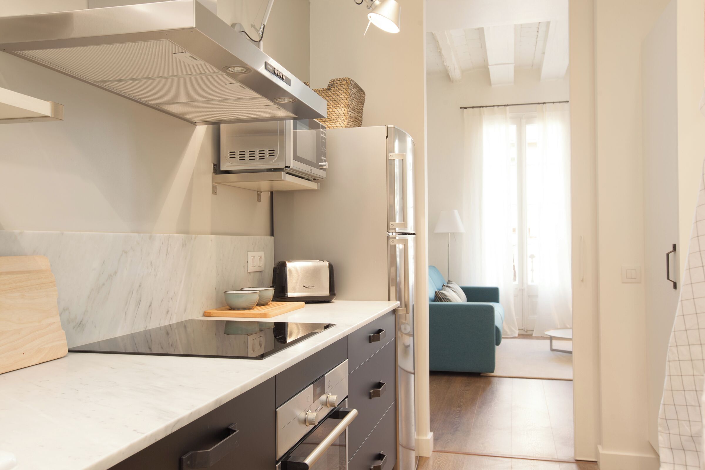 Una cocina moderna con microondas, tostadora y refrigerador. Hay una tabla de cortar de madera y un bol en la encimera. La cocina tiene un protector contra salpicaduras blanco y armarios grises.