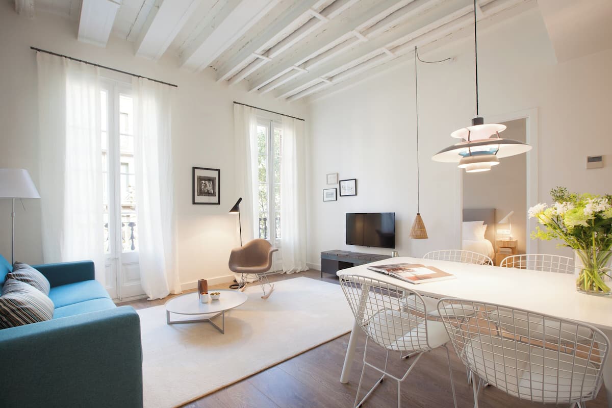 Una sala de estar moderna con un sofá, una mesa de comedor con sillas, un televisor y un ventanal con cortinas.