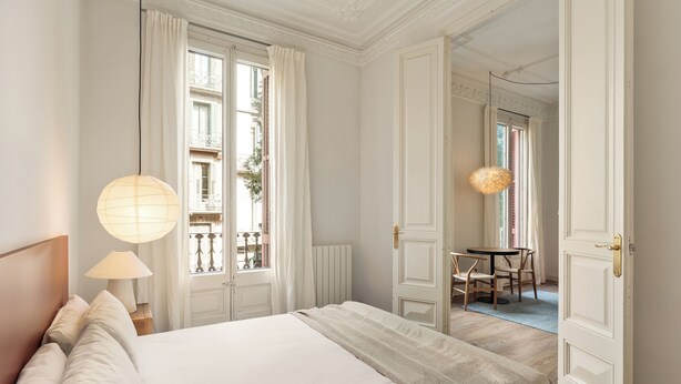 A hotel room with a large bed, a bedside table with a lamp, a window with curtains, and a door leading to another room. - En Casa de Girona 17 (Barcelona)