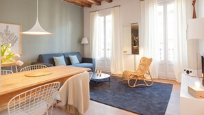 A modern living room with a wooden ceiling, a blue sofa, a dining table with chairs, a wicker rocking chair, and a TV on a white stand. - En Casa de Girona 17 (Barcelona)