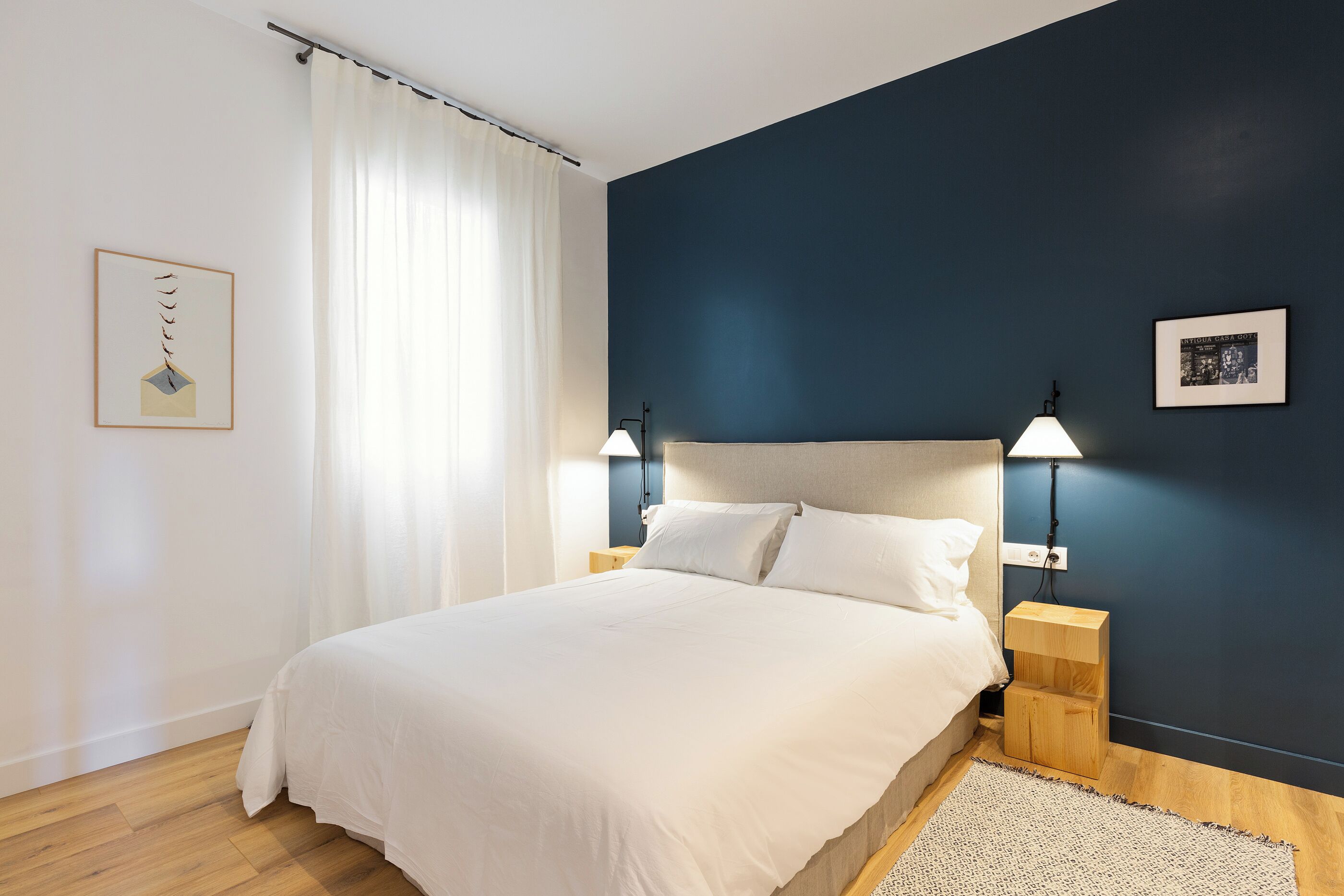 A bedroom with a bed, two bedside tables, a nightstand, a wall-mounted lamp, and a framed picture on the wall.