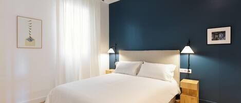 A bedroom with a bed, two bedside tables, a nightstand, a wall-mounted lamp, and a framed picture on the wall.