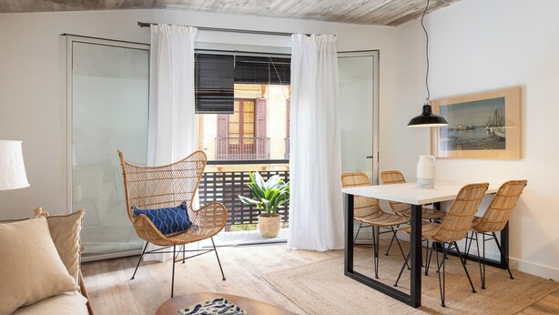 Un espacio habitable moderno con sofá, mesa de comedor, sillas de mimbre, una planta en maceta y vistas a un balcón con barandilla y un edificio al fondo.