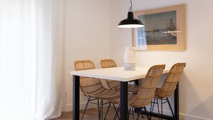 Un comedor con una mesa blanca, sillas de mimbre, una lámpara colgante negra y un cuadro en la pared.