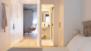Un dormitorio con cama, mesita de noche y tocador. Se ve un baño con lavamanos y espejo. Una silla está colocada en una esquina de la habitación.