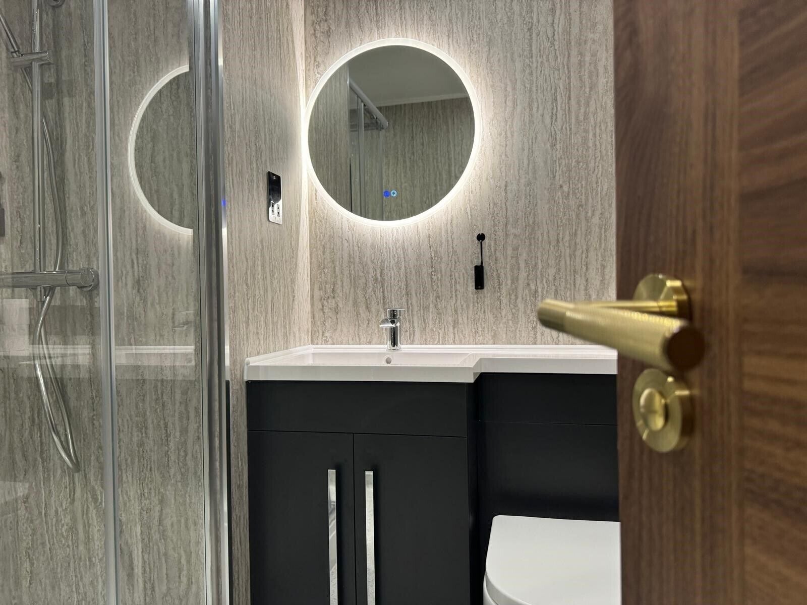 Une salle de bain moderne avec une douche à l’italienne en verre, un miroir rond et un lavabo blanc.