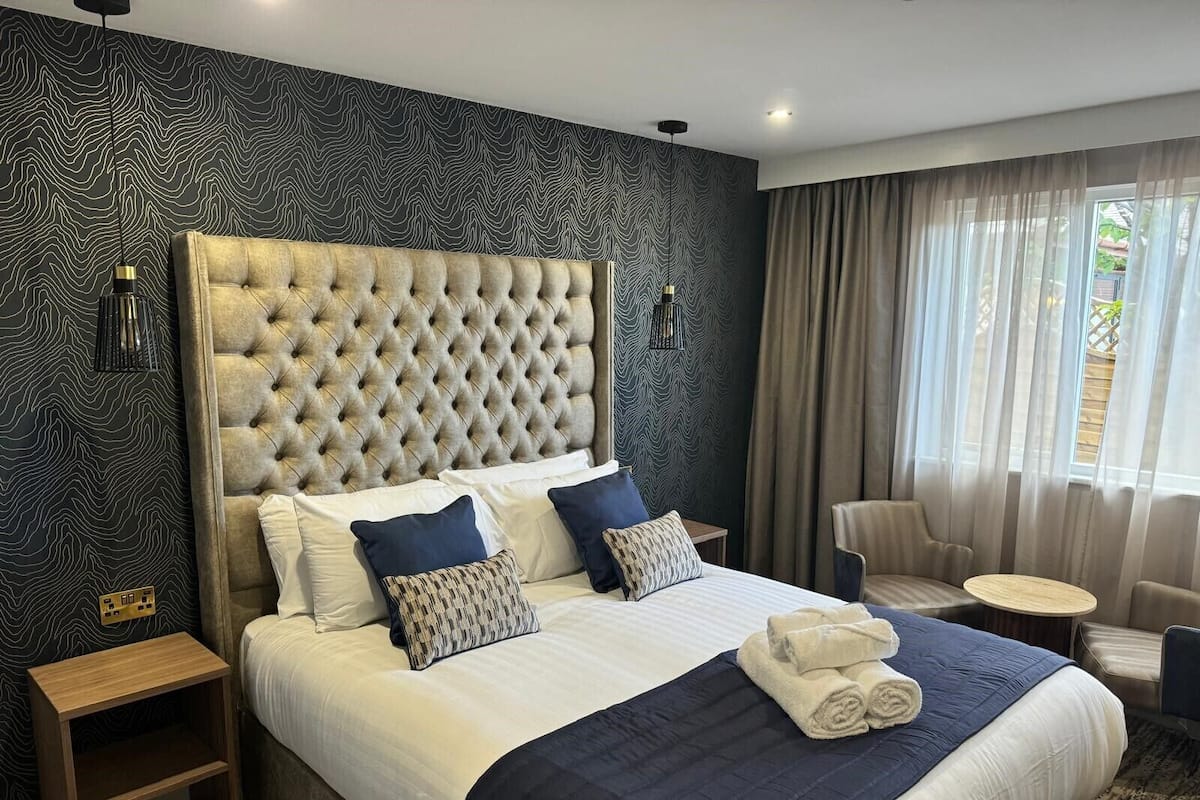 Ein Hotelzimmer mit einem großen Bett, einem Kopfteil, einem Nachttisch, einem Sessel, einem runden Tisch und einem Fenster mit Vorhängen.