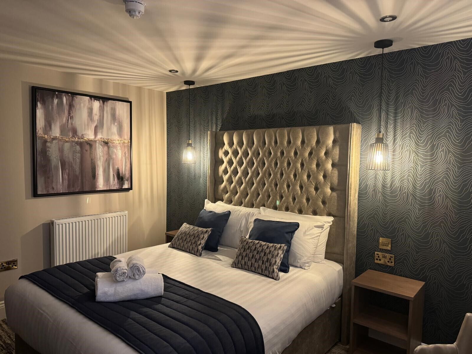 A modern hotel room with a large bed, a headboard with a diamond pattern, and a textured wall.