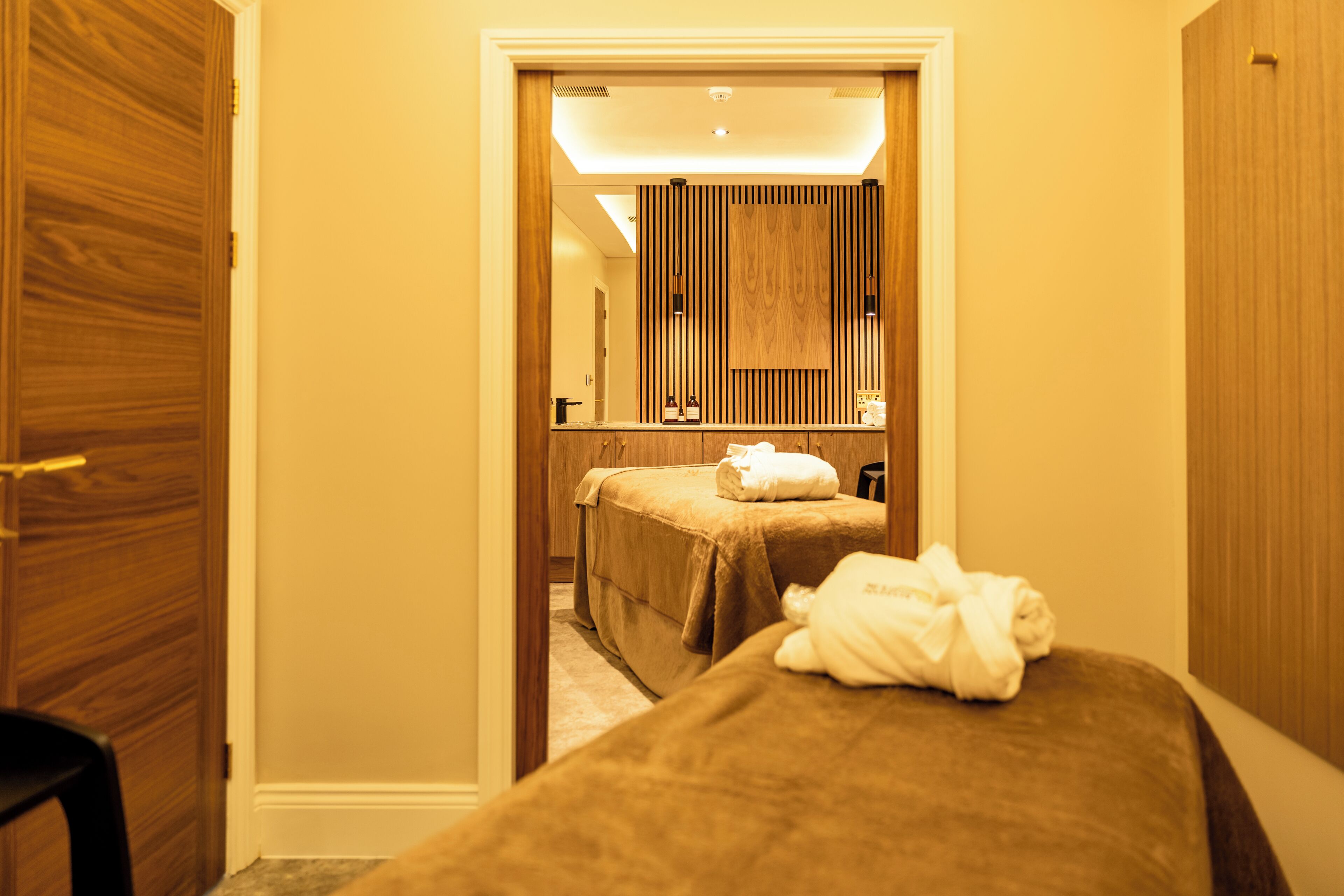 A hotel room with a wooden wardrobe, a bed with a brown bedspread, a small table with a bottle and a glass, and a mirror above the bed.