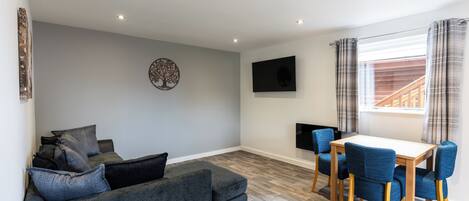 A modern living room with a grey sofa, a wooden dining table with blue chairs, a flat-screen TV mounted on the wall, and a window with curtains.
