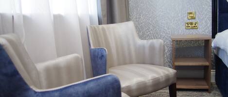 A hotel room with two armchairs, a wooden nightstand, and a bed.