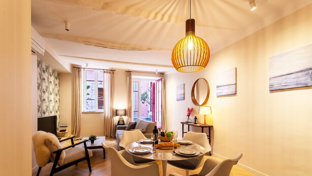A dining area with a round table, white chairs, a hanging light fixture, and a view of the living room through an open doorway.