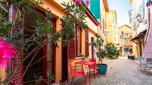 A cobblestone street with a café featuring red chairs and a table, surrounded by colorful buildings with shutters.