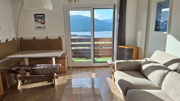 A modern living room with a wooden table, a sofa, and a bench. A large window offers a view of a mountainous landscape.