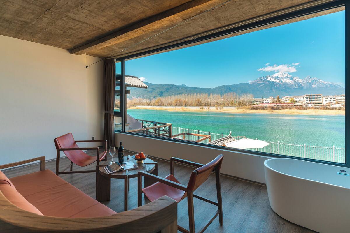 Ein Balkon mit Blick auf einen See, Berge und einen Strand. Zwei Stühle und ein Tisch mit einer Flasche und Gläsern.