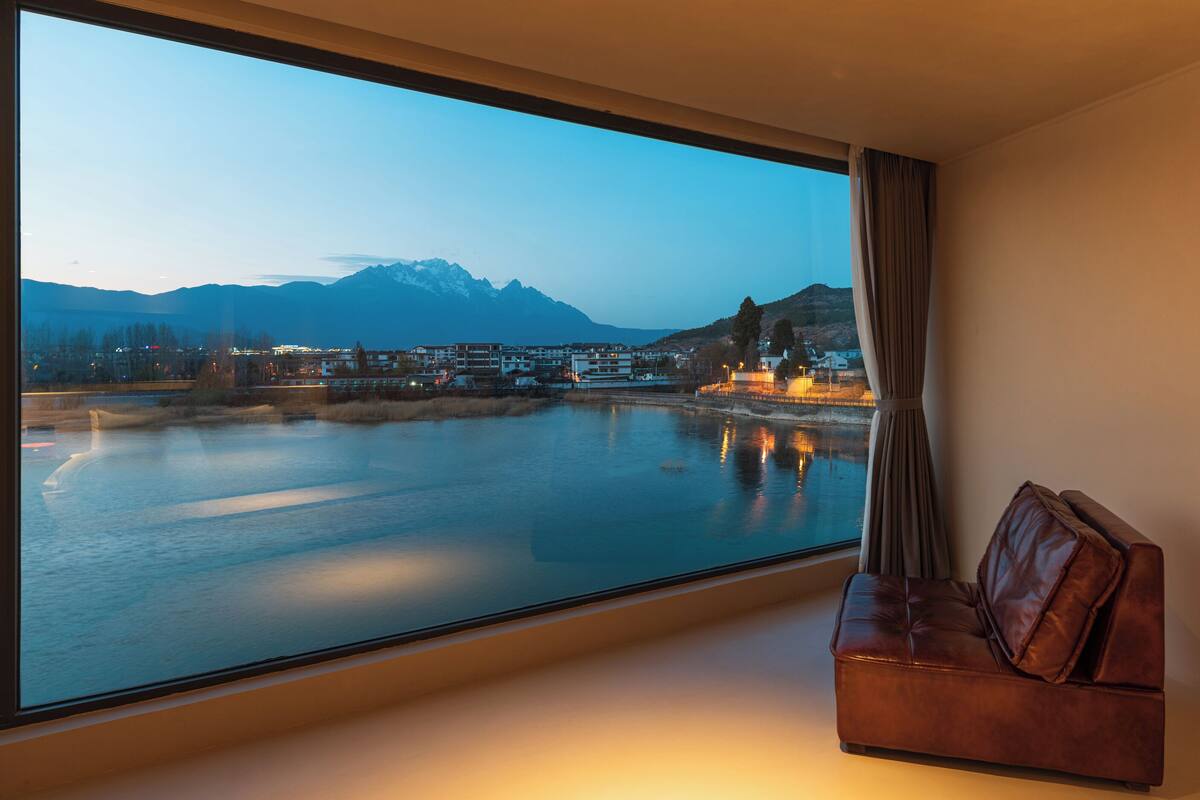 Ein Zimmer mit großem Fenster mit Blick auf einen ruhigen Fluss und Berge, ein Ledersofa und ein Vorhang.